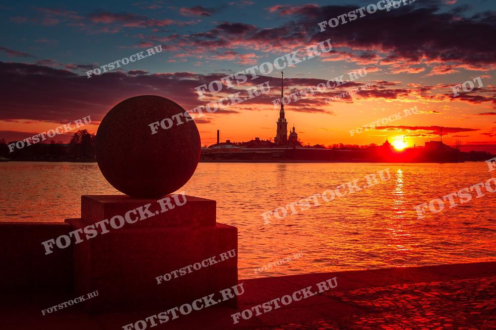 На фотографии запечатлен восход солнца над рекой Невой в Санкт-Петербурге с видом на Петропавловскую крепость. Силуэт гранитного шара на переднем плане добавляет композиции глубину и интерес. Небо окутано волшебными оттенками оранжевого и красного цветов, которые отражаются на водной глади. Такая фотография может быть использована в туристических брошюрах, для дизайна обложек книг и журналов, в арт-проектах, а также как элемент оформления интерьера.